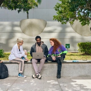 Three people sit outdoors on a campus bench, collaborating while viewing a laptop.